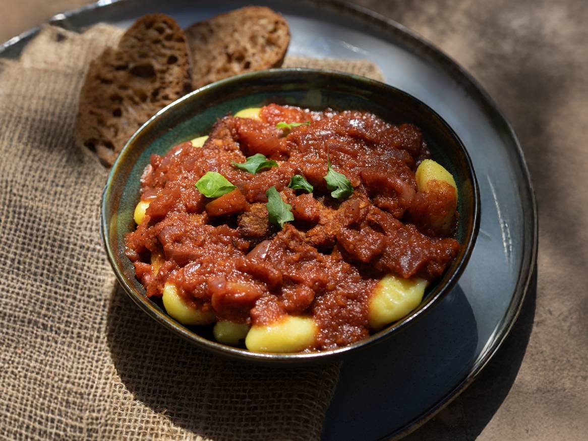 Njoki s gulašem od rajčice i govedine, posluženi s kruhom na rustikalnoj podlozi.