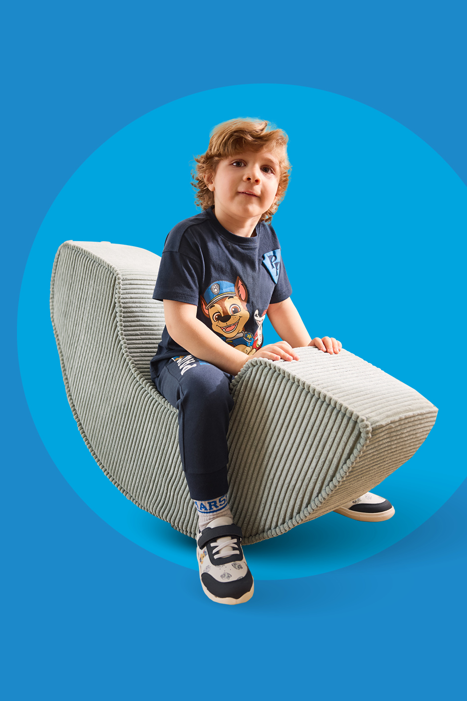 Dječak u tamnoplavoj majici s printom i trenirci sjedi na sivoj fotelji.