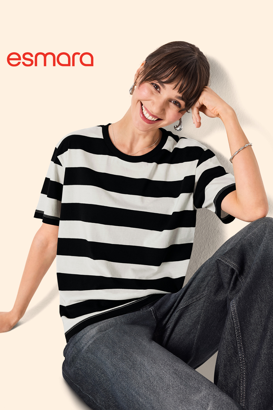 Žena u crno-bijeloj prugastoj majici i tamnim trapericama, nasmijana, naslonjena na zid.