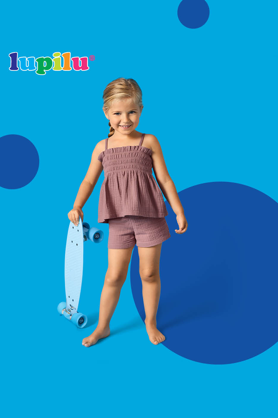 Djevojčica u ružičastom kompletu s topom i kratkim hlačama drži plavi skateboard.