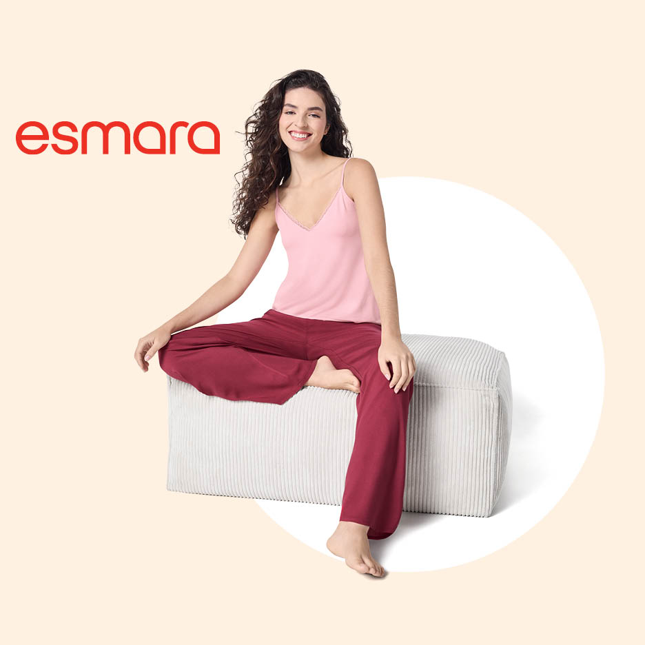 Žena u ružičastoj majici na naramenice i širokim bordo hlačama sjedi na tabureu.