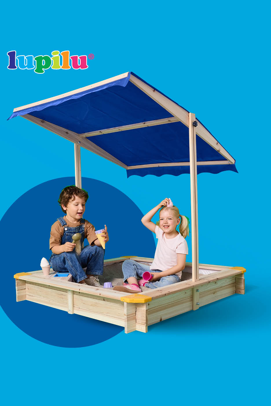 Dvoje djece igra se u pješčaniku s plavim krovom, dječak drži lopaticu, a djevojčica se smije.