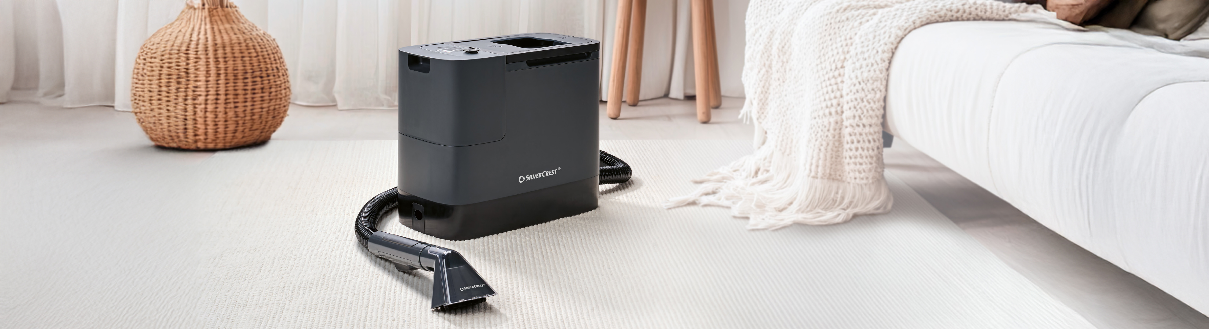 Sivi uređaj za čišćenje tepiha s crijevom i mlaznicom na svijetlom tepihu u dnevnoj sobi.