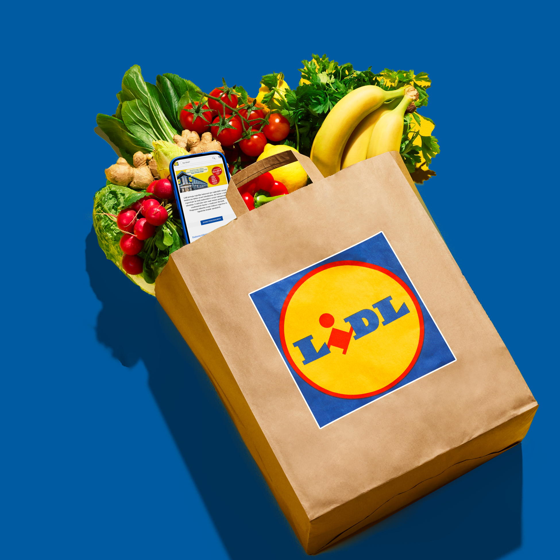 Papirnata vrećica s namirnicama, uključujući banane, rajčice, rotkvice i zeleno lisnato povrće, s telefonom koji prikazuje aplikaciju za kupovinu.