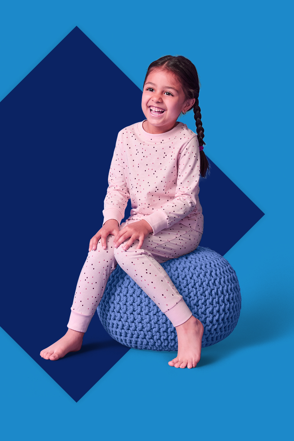 Djevojčica u ružičastoj pidžami s točkicama sjedi na plavom pufu i smije se.