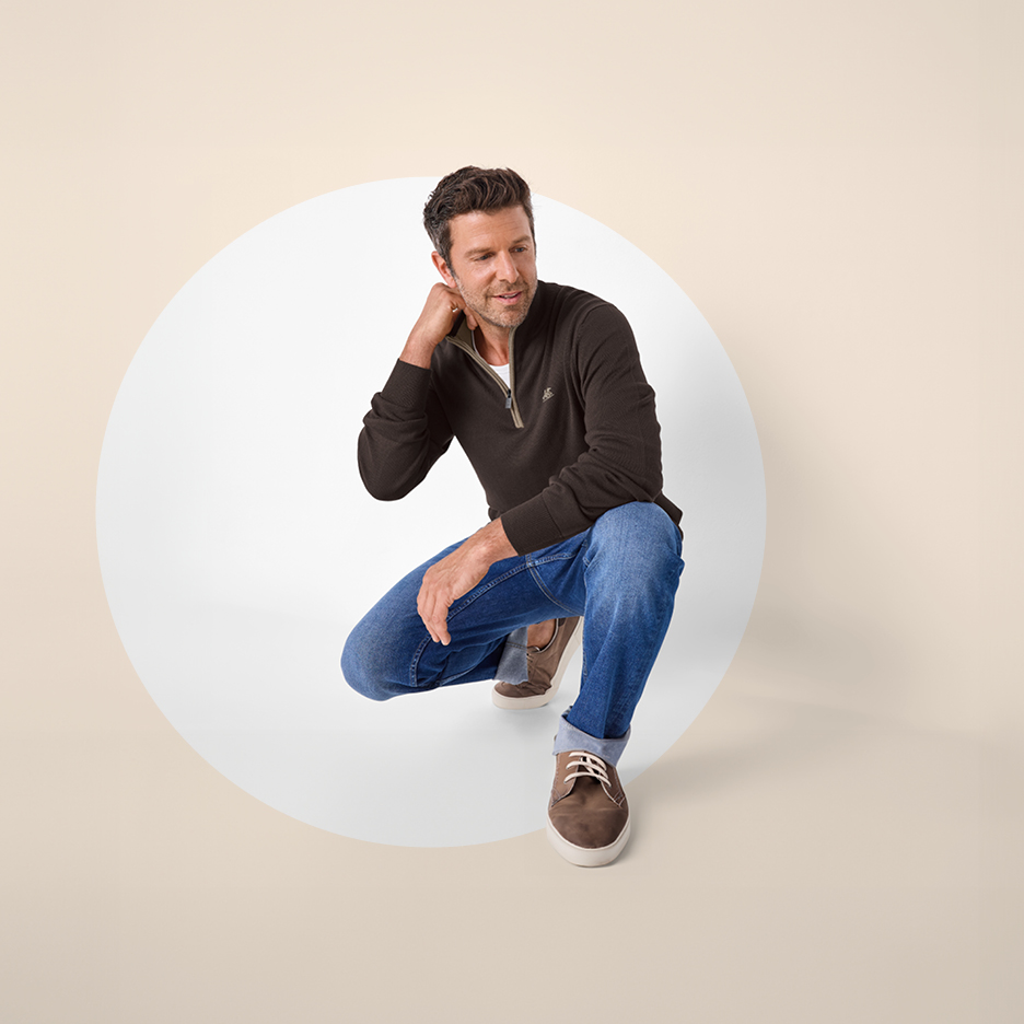 Muškarac u tamnosmeđoj pletenoj majici s patentnim zatvaračem i trapericama čuči u bijelom krugu.